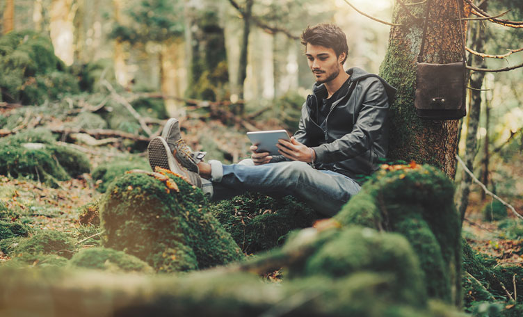 Un joven leyendo con una tablet apoyado en un árbol en un bosque